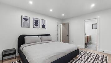 A modern bedroom with a large bed, a bedside table, and a nightstand. There are three framed artworks on the wall and a door leading to another room.