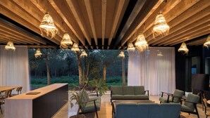 Un interior moderno con techo de madera, lámparas colgantes, mostrador de recepción, zona de estar y vistas a árboles.