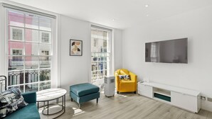 Een moderne woonkamer met een bank, een poef en een gele fauteuil. Een flatscreen televisie hangt aan de muur, en eronder staat een witte tv-meubel.