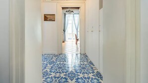A hallway with patterned floor tiles, a chandelier, and a view into a room with a large window and curtains. - Ammore A Mare - Luxury Apartment Near The Sea (Napoli)