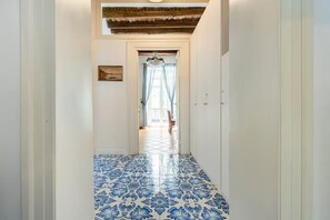 Un couloir avec des carreaux de sol à motifs, un lustre et une vue sur une pièce dotée d’une grande fenêtre et de rideaux.