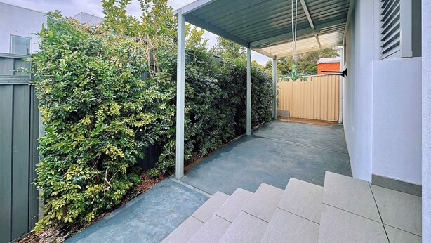 A covered outdoor area with a concrete pathway leading to a wooden door, surrounded by dense green foliage. - 1BR Granny Flat / Free Parking / Near Airport & Train (Arncliffe)