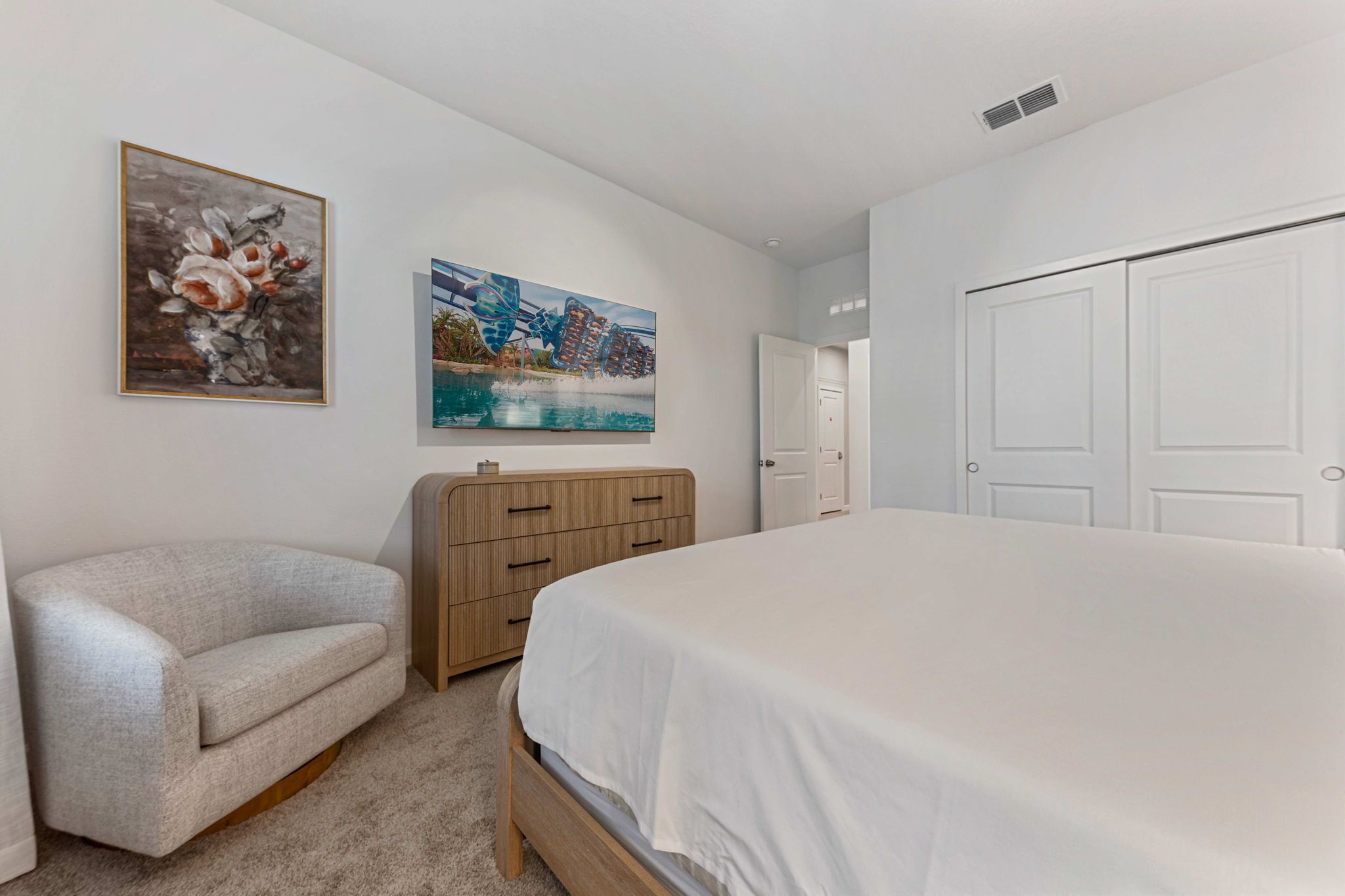 A bedroom with a bed, a wooden dresser, a gray armchair, and two paintings on the wall.