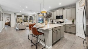 A modern kitchen with a central island, bar stools, pendant lighting, and a dining area with a sofa and coffee table. - 2332 Distant Sun Trail (Kissimmee)