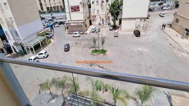 Une vue depuis un balcon donnant sur un parking où se trouvent plusieurs voitures, quelques arbres et, au loin, des bâtiments.