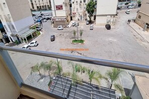 A view from a balcony overlooking a parking lot with several cars, a few trees, and buildings in the background. - Warm, modern apartment in the heart of Akid Lotfi (Oran)