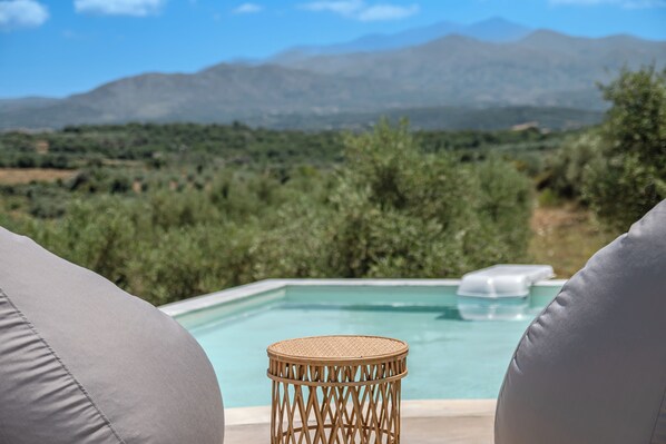 A poolside view with a wicker table and two grey lounge chairs, overlooking a mountainous landscape.