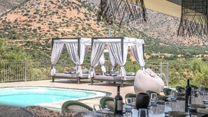 Zona junto a la piscina con vistas a un paisaje montañoso, mobiliario exterior con toldos y una mesa lista para comer.