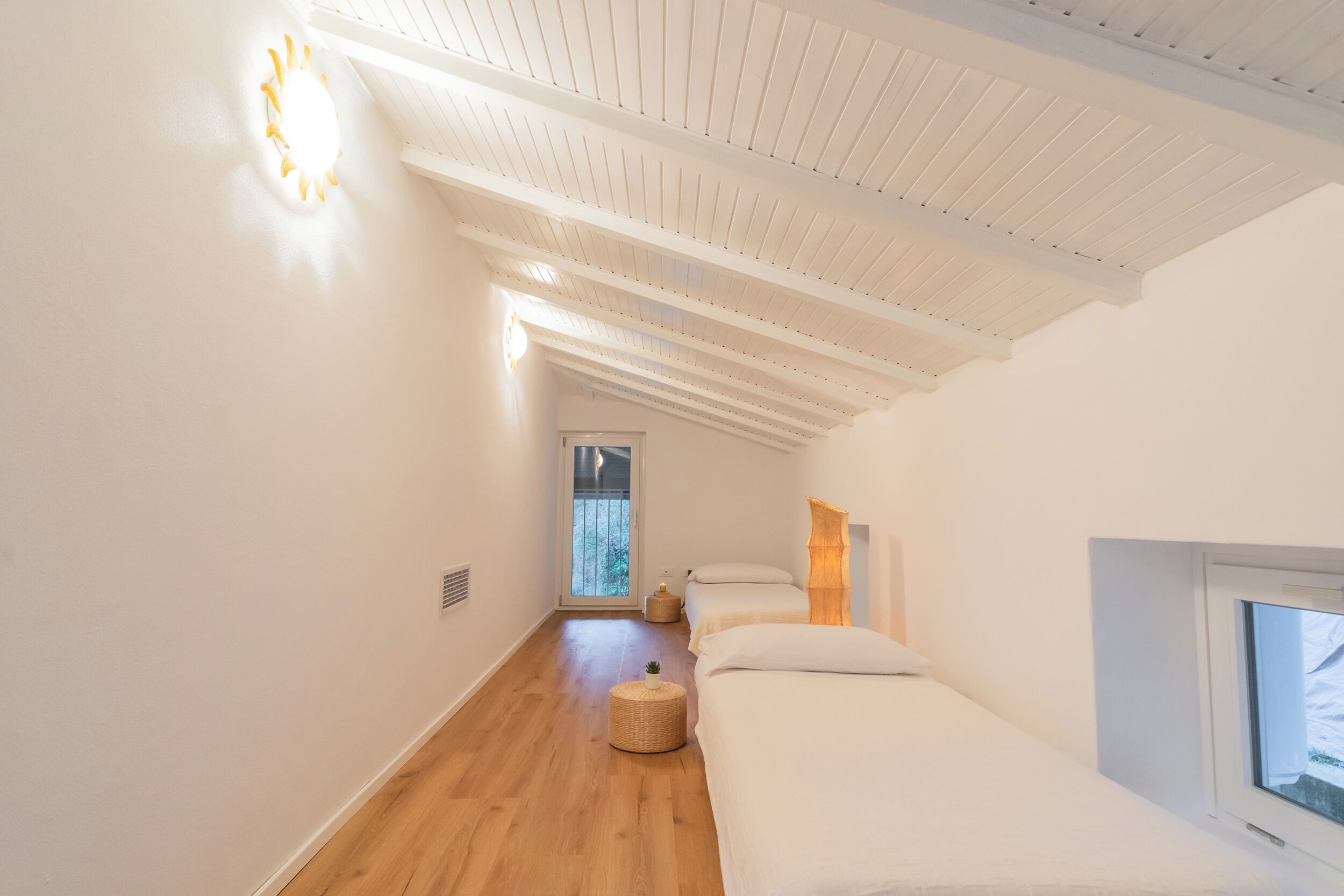 A narrow hallway with wooden flooring leading to a bedroom with two beds and a window.