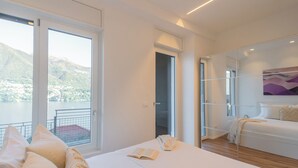 A modern bedroom with a large bed, a bedside table, and a view of a lake. - Villa Regina Retreat With Pool & Lake View (Brienno)