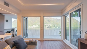 A modern living room with a large sliding glass door leading to a balcony with a mountain view. - Villa Regina Retreat With Pool & Lake View (Brienno)