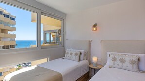 Hotellihuone, jossa on kaksi sänkyä, suuri ikkuna, josta on näkymä merelle ja rakennuksiin, sekä pieni pöytä, jossa on lamppu.