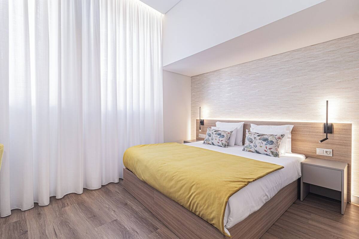 A modern hotel room with a wooden bed, white curtains, and a nightstand with a lamp.
