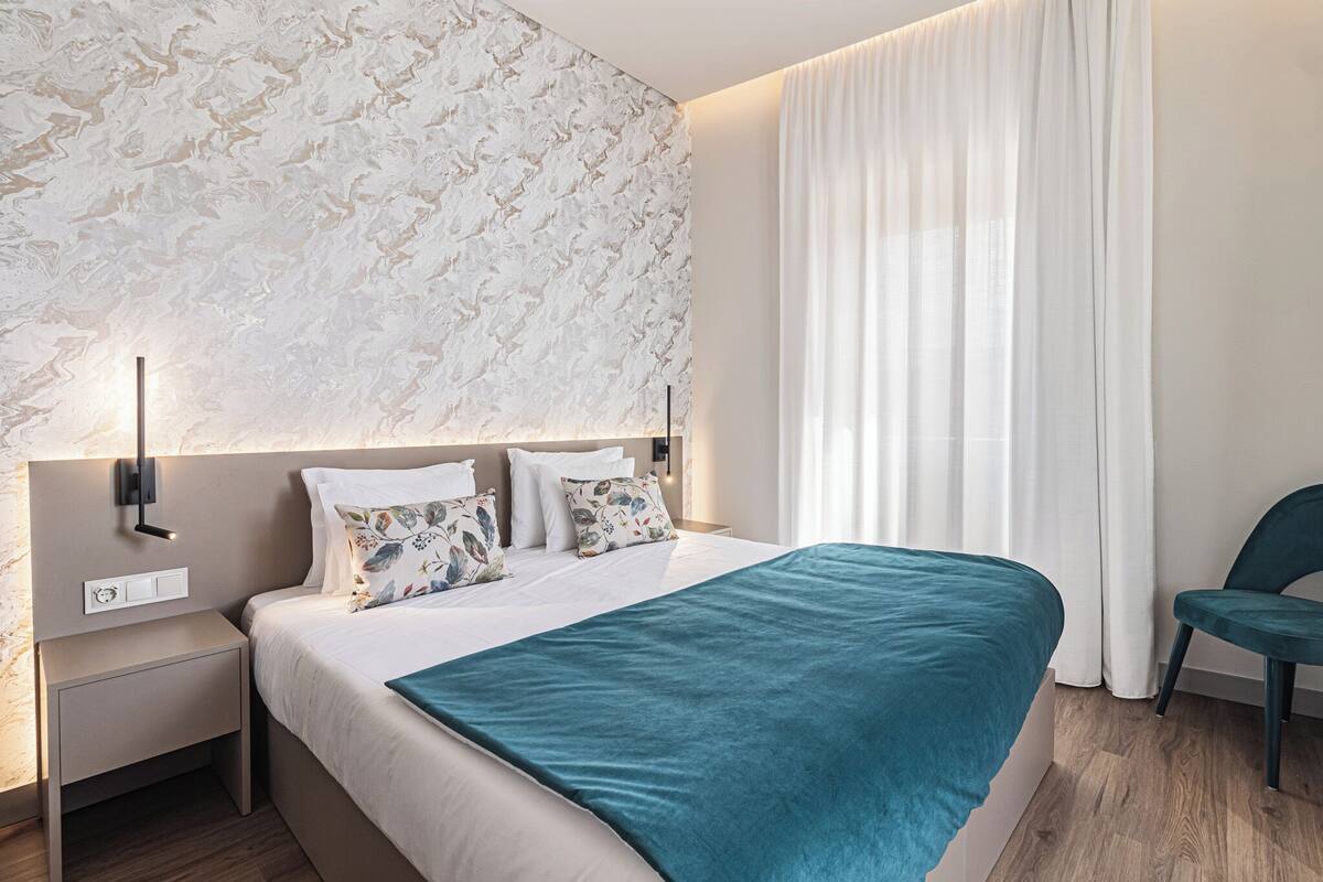 A modern hotel room with a large bed, a nightstand, a wall-mounted lamp, and a window with curtains.