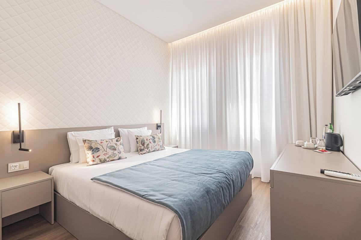 A modern hotel room with a neatly made bed, a bedside table with a lamp, and a large window with curtains.