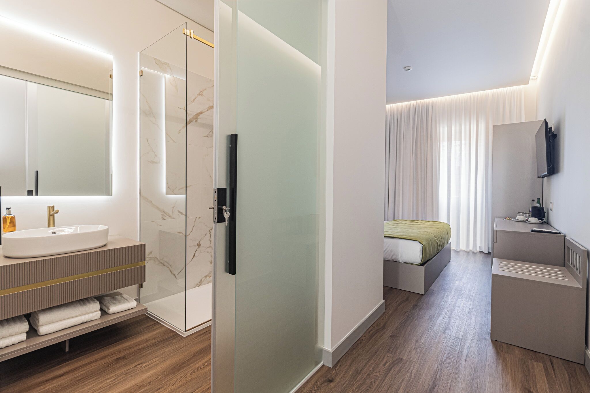 Una habitación de hotel moderna con tocador de mármol, cabina de ducha de vidrio, cama con cubrecama verde y televisor de pantalla plana montado en la pared.