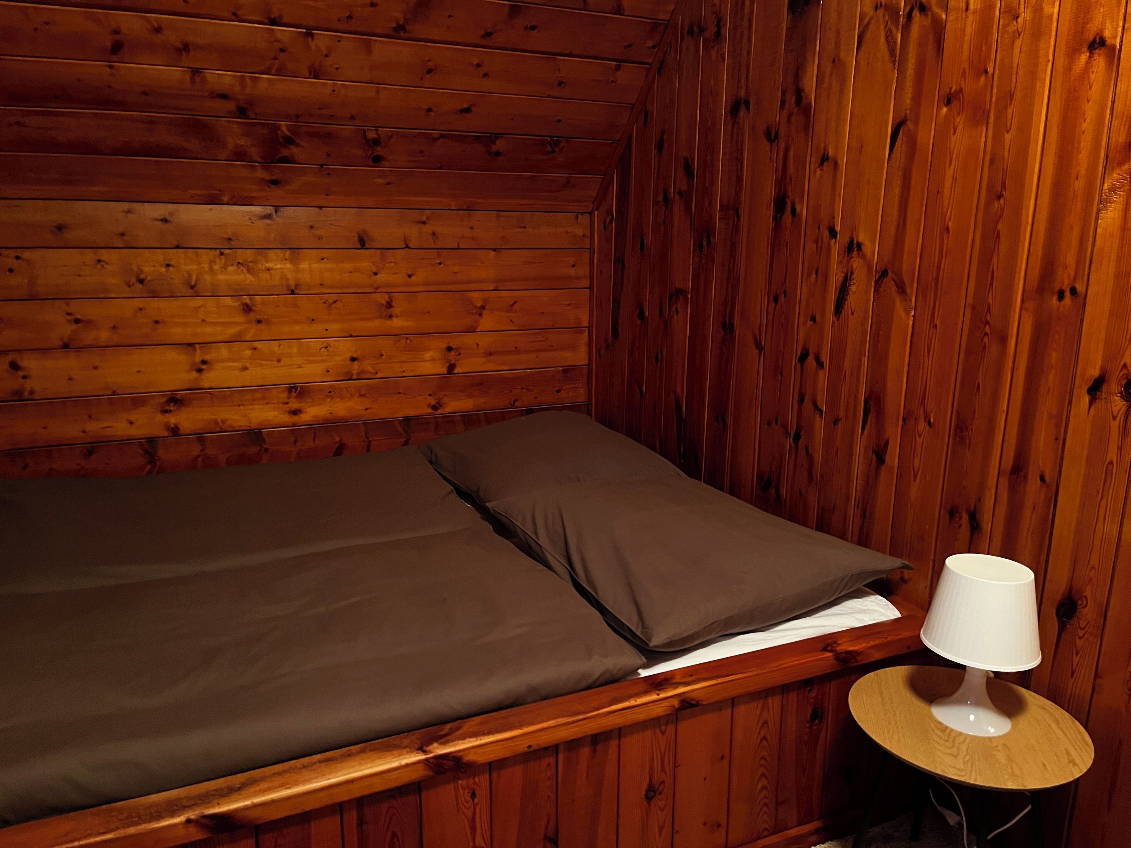 A wooden cabin room with a single bed, a small round table with a lamp, and a wooden wall.