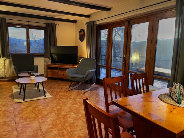 A cozy living room with a TV, a grey armchair, a wooden dining table, and a view of a snowy landscape through large windows.