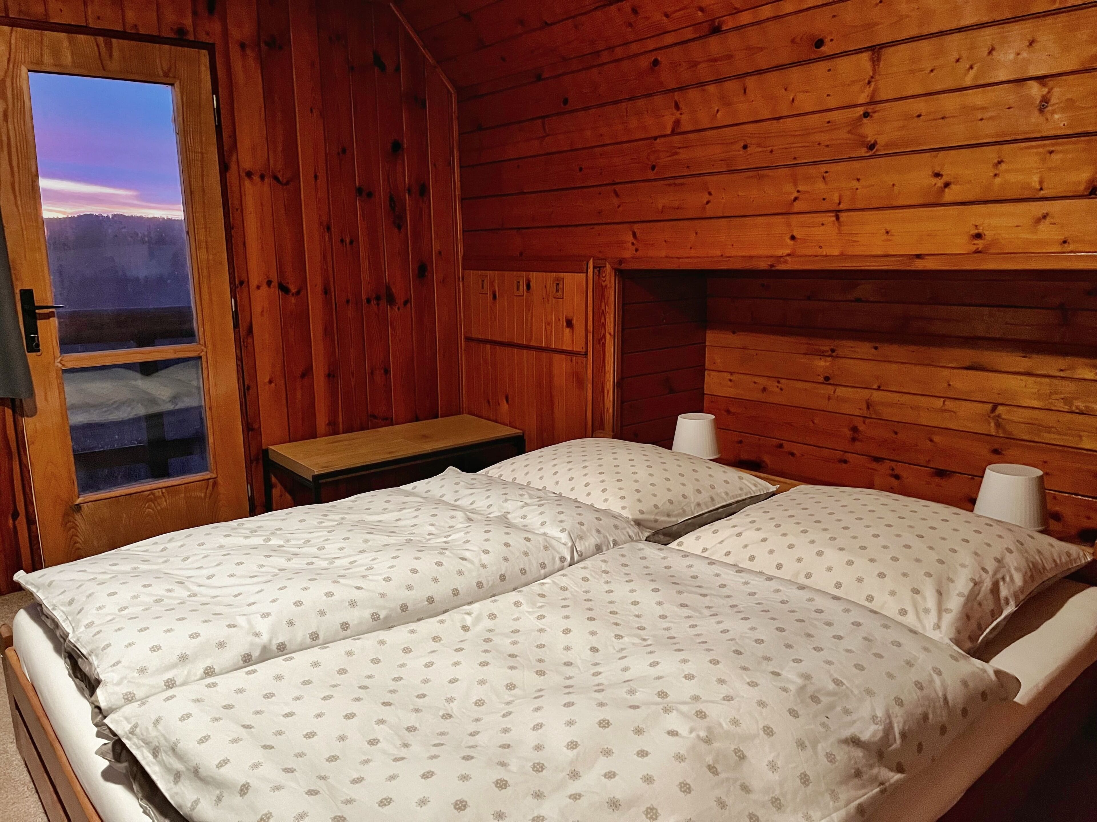 A wooden cabin room with a bed, a nightstand, and a window.