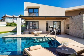 Un área de piscina moderna con una silla de descanso, una mesa con bebidas y vistas a la casa.