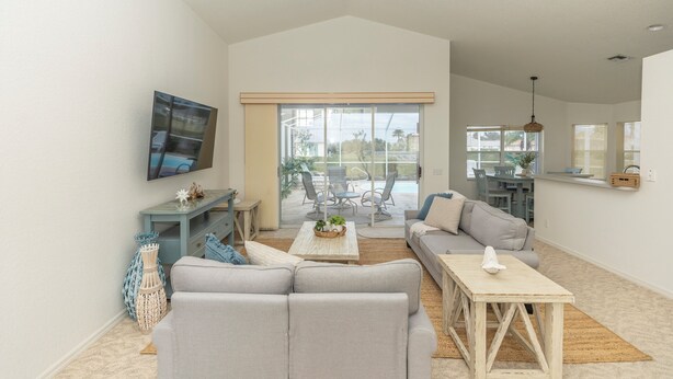A spacious living room with a large sofa, a coffee table, and a TV mounted on the wall. There is a sliding glass door leading to an outdoor patio area. - Rotonda Coastal 3 bed 2.5 bath stunning pool home next to golf course! (Rotonda West)