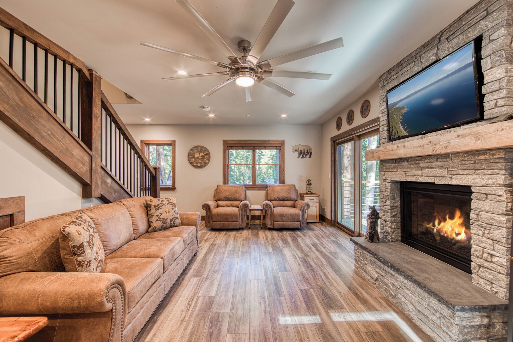 A spacious living room with a stone fireplace, a large flat-screen TV mounted above it, a ceiling fan, and a wooden staircase leading to another level.