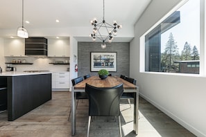 Een moderne keuken met een centraal eiland, een eethoek met een houten tafel en zwarte stoelen, en een groot raam met uitzicht op bomen.