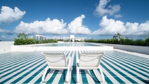 A pool area with two white lounge chairs facing a pool, surrounded by a blue and white tiled deck, under a clear sky.