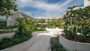 A courtyard with a paved walkway, surrounded by planters with various plants and trees, and a building with balconies in the background.