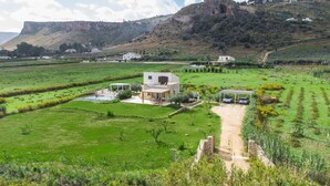 A modern house with a large patio, surrounded by lush green fields and a mountain backdrop. - Casa Maruzza | Scopello | Sea-view retreat with infinity pool (Castellammare del Golfo)