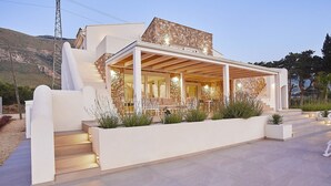 Een modern huis met een gevel van natuursteen, een overdekte patio met buitenmeubels en een trap die naar de ingang leidt.