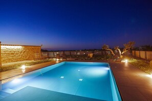 Ein rechteckiger Pool mit einer Steinwand an einer Seite und einem Holzzaun an der anderen, umgeben von einem gut beleuchteten Terrassenbereich.