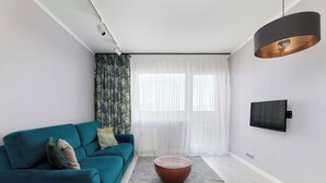 Una sala de estar moderna con un sofá color turquesa, una mesa de centro redonda de madera, un televisor montado en la pared y cortinas translúcidas.