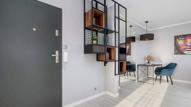 Una habitación moderna con una puerta oscura, un estante montado en la pared con varios objetos, una pequeña mesa con una silla y un cuadro colorido en la pared.