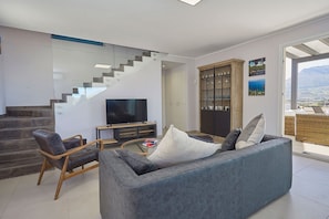 A modern living room with a grey sofa, wooden chair, and a flat-screen TV. A staircase leads to a balcony with a view of the landscape. - Casa Fior di Mandorla | Castellammare del Golfo | Sea View | Infinity Pool (Castellammare del Golfo)