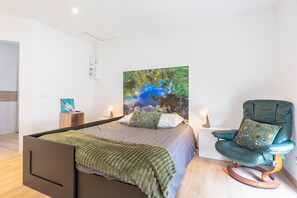 Un dormitorio con cama, una mecedora, un cuadro en la pared y una mesita de noche con lámpara.