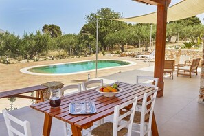 Ein Essbereich im Freien mit Pool, Holztisch, weißen Stühlen und einer Steinwand.
