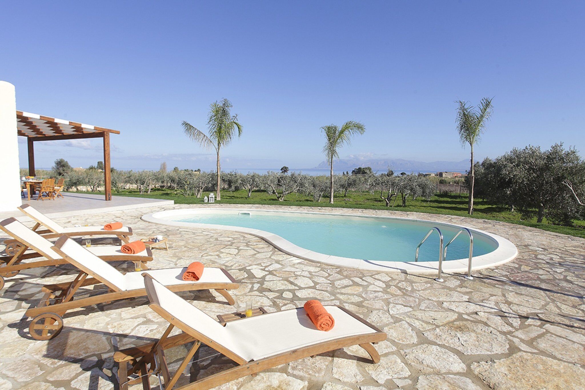 Un espace piscine agrémenté de chaises longues et d’une pergola, entouré d’oliviers et offrant une vue dégagée sur la mer.