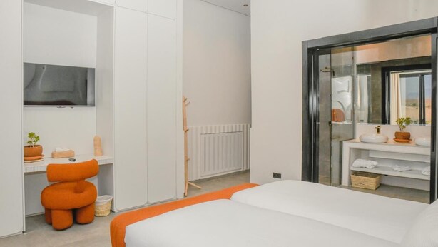 Ein modernes Hotelzimmer mit einem Bett, einem an der Wand montierten Fernseher, einem Schreibtisch mit Stuhl sowie einem Badezimmer mit Spiegel und Waschbecken.