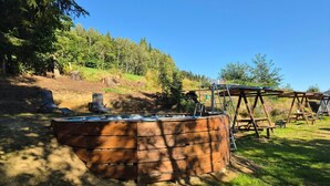 Entorn rural a l'aire lliure amb una estructura de fusta, possiblement una banyera d'hidromassatge, envoltat per un paisatge natural amb arbres i un cel serè.