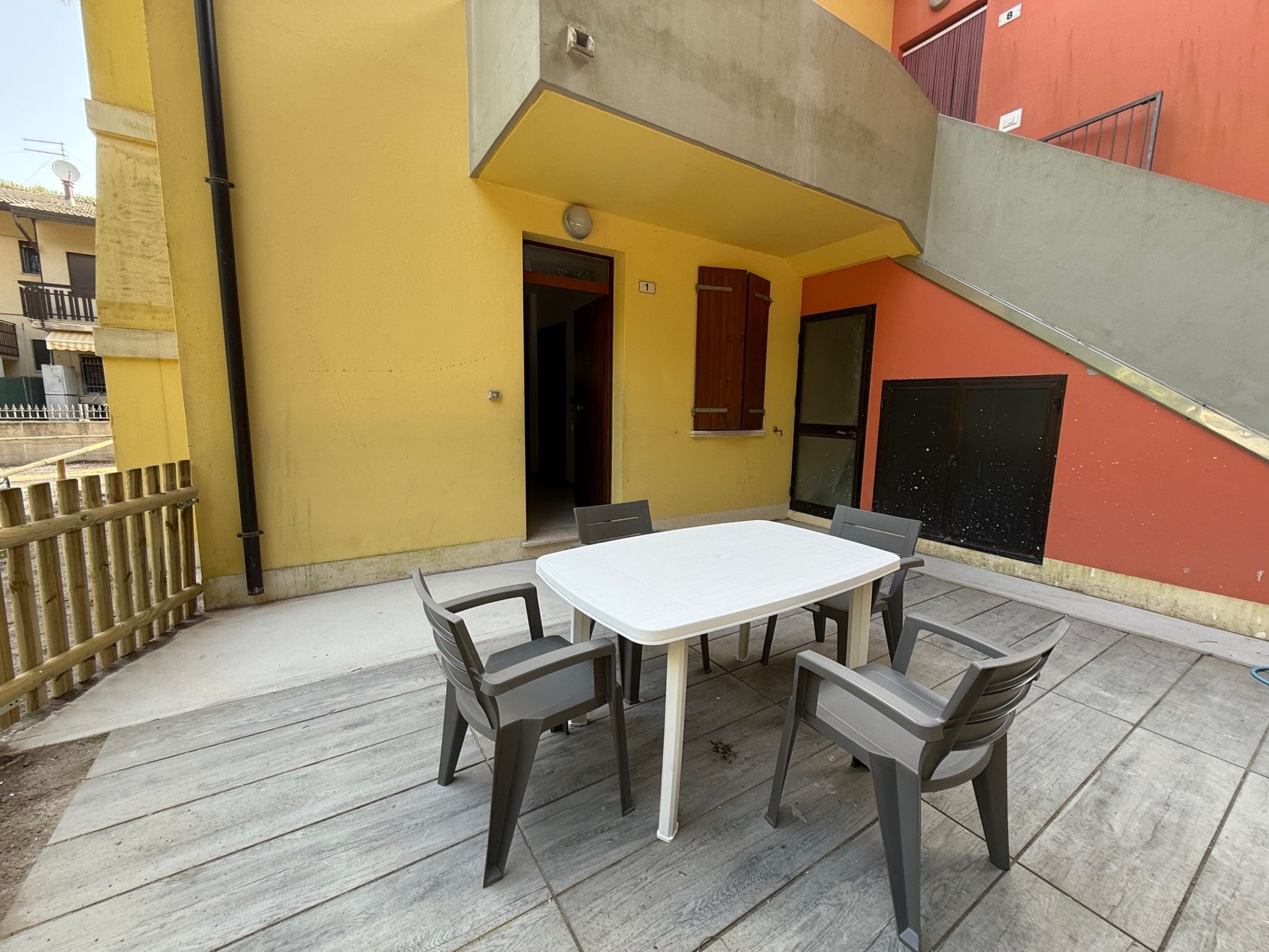 Eine Terrasse mit einem weißen Tisch und vier Stühlen, einem Holzzaun und einem gelben Gebäude mit rotem Abschnitt.