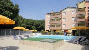Una piscina con lettini e ombrelloni gialli, circondata da un solarium piastrellato e un edificio a più piani con balconi.