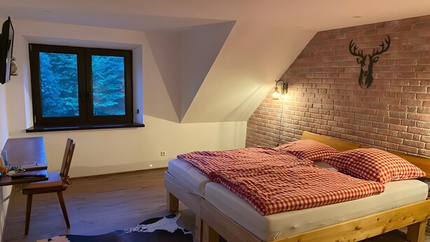 A bedroom with a sloped ceiling, a bed with a checkered blanket, a wooden desk, a laptop, a chair, a cowhide rug, and a brick wall with a deer head decoration.