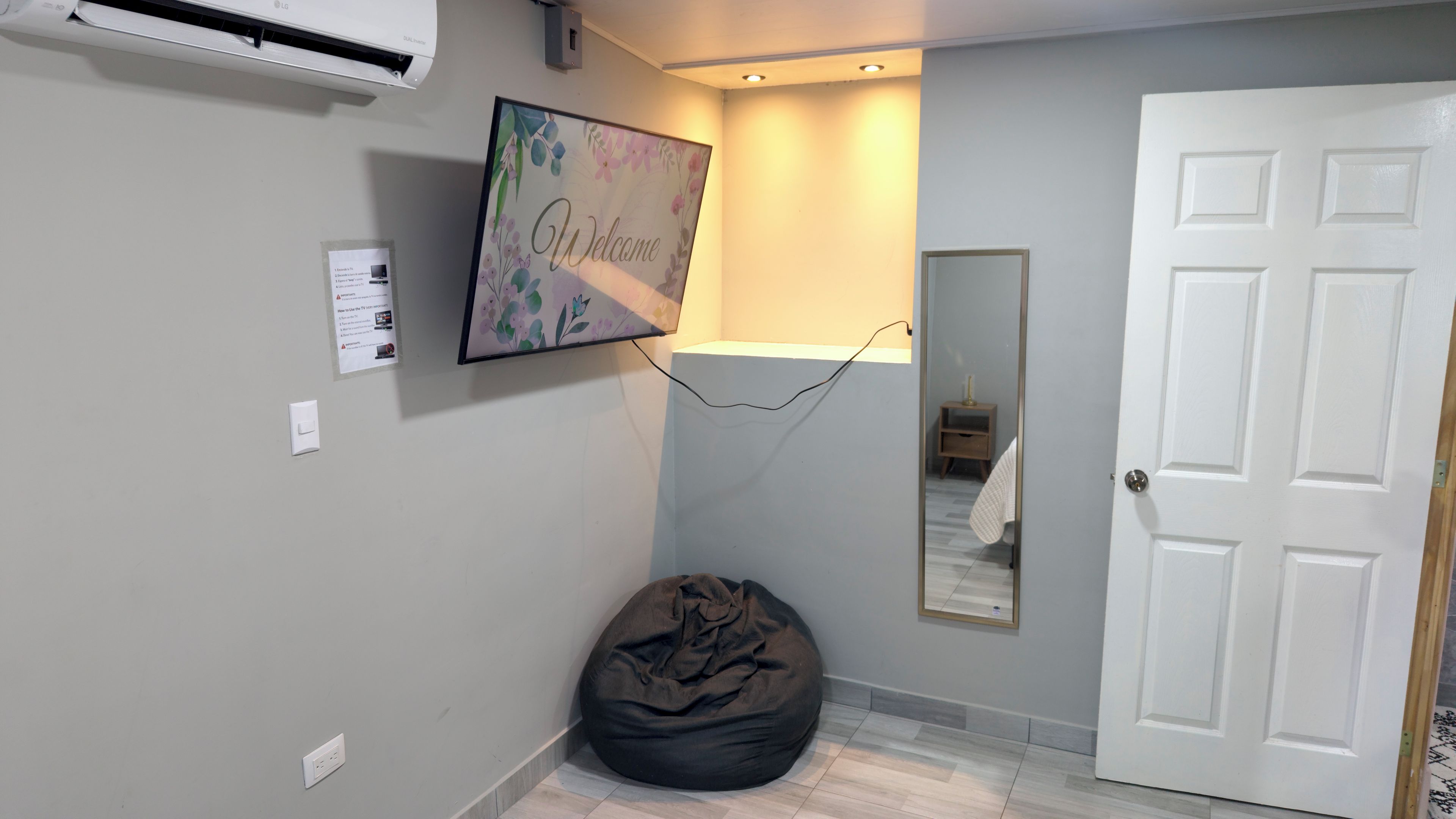 Un couloir avec une porte blanche, un fauteuil en forme de sac de haricots noir, un téléviseur fixé au mur et un panneau d’accueil.