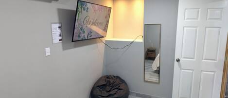 Un passadís amb una porta blanca, un seient informal negre, un televisor penjat a la paret i un cartell de benvinguda.