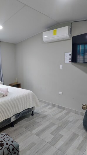Habitació d'hotel amb un llit, aire condicionat, una taula de nit, un televisor fixat a la paret i un otomà estampat.