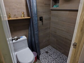A small bathroom with a toilet, a shelf with decorative items, a shower curtain, and a tiled floor.