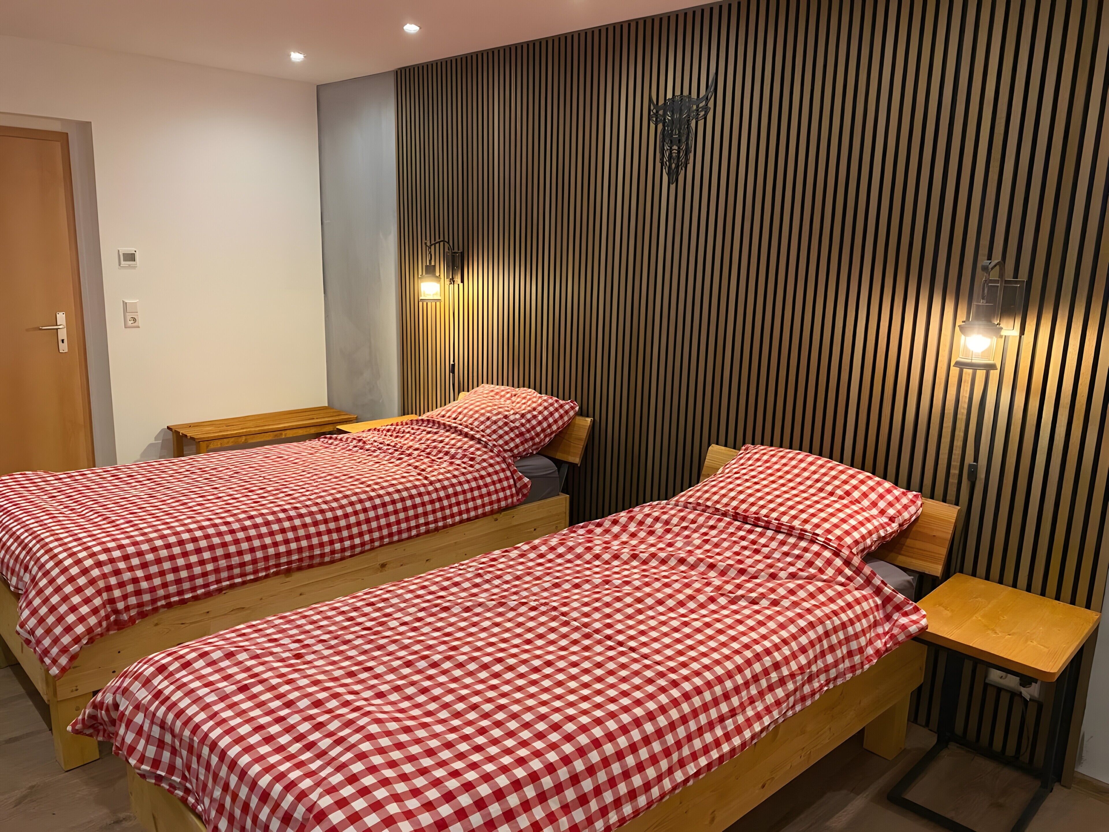 A room with two wooden beds, a bench, a small table, and a wall-mounted light fixture.