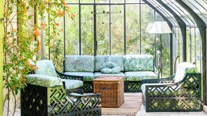 Ein Wintergarten mit einer grünen Sofagarnitur, einem Korbstuhlhocker und Gitterstühlen.