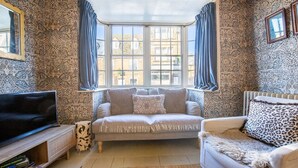 Uma sala de estar aconchegante com um parapeito, papel de parede estampado, um sofá, uma poltrona com estampa de leopardo e uma televisão sobre um suporte de madeira.